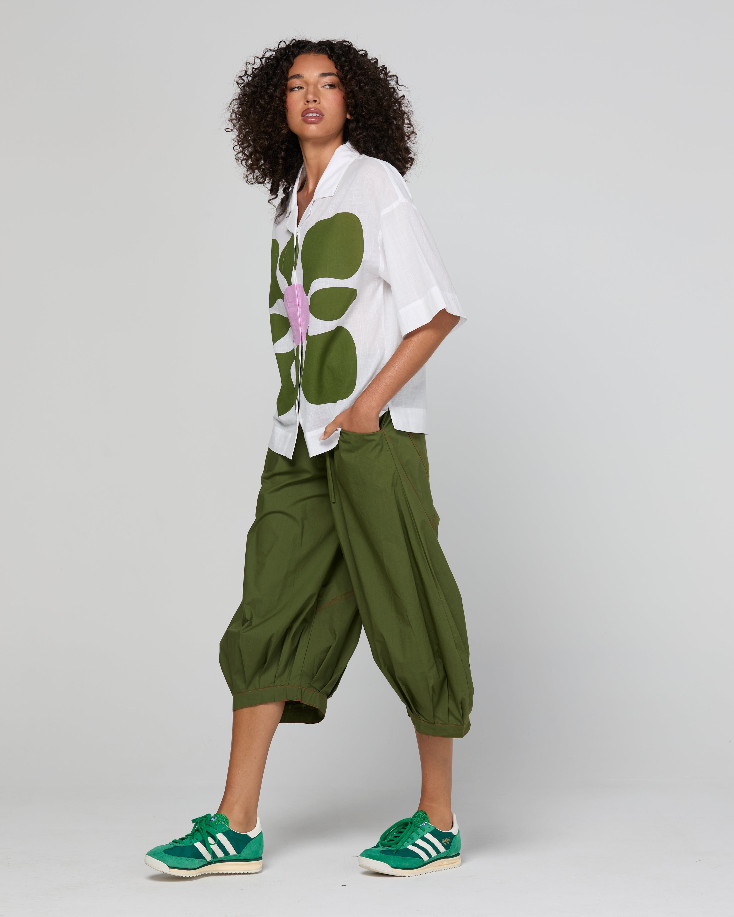 Woman wearing a white shirt with a green floral pattern and green pants on a plain background