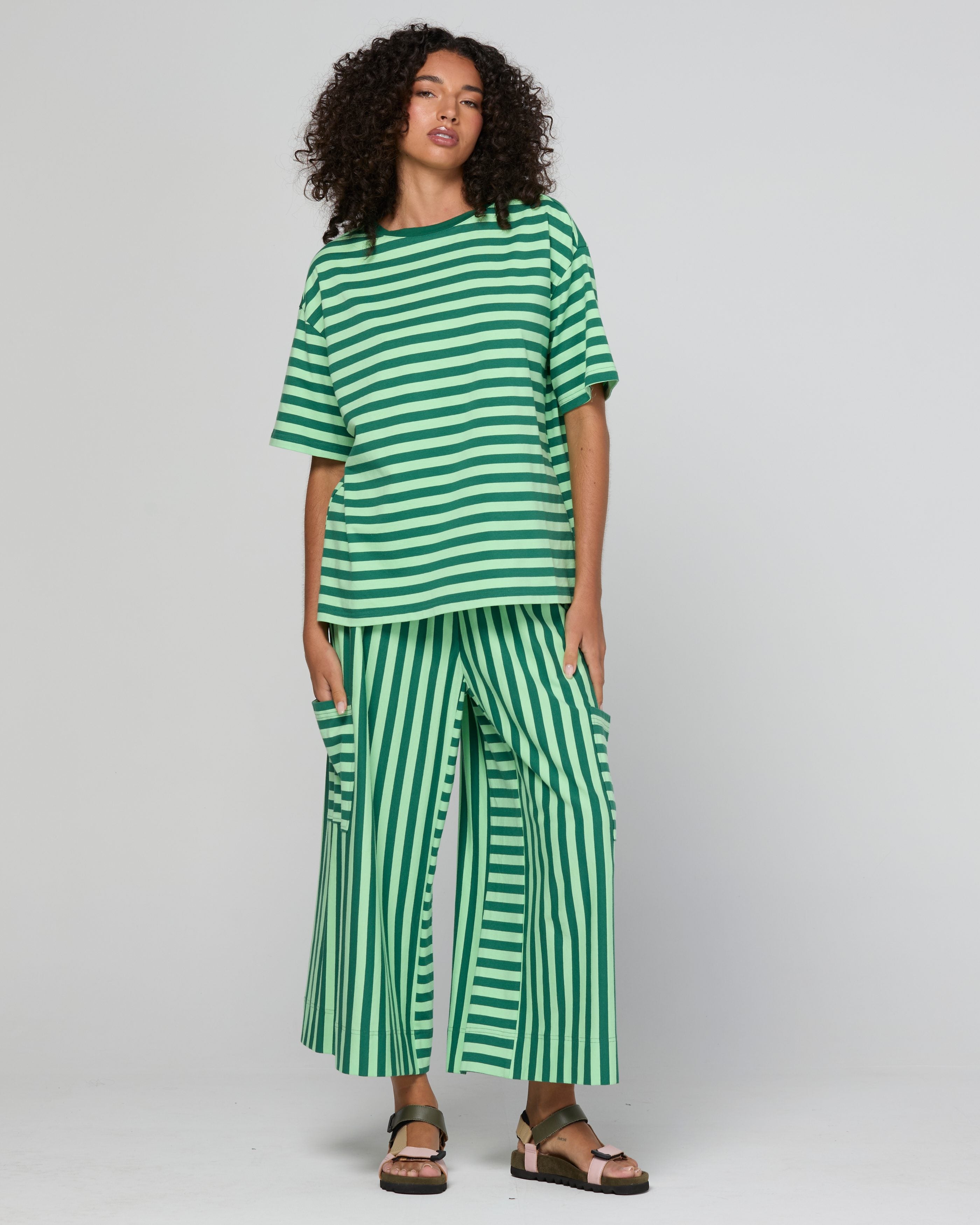 A model wearing a green and white striped two-piece set consisting of a short-sleeve top and wide-leg pants, paired with brown sandals, against a light gray studio background.