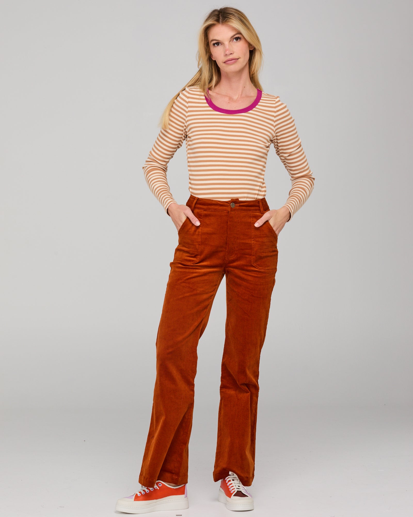 Woman wearing a cream striped shirt and rust corduroy pants on a gray background