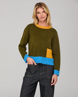 Woman wearing an olive jumper with color-blocked design, and dark denim jeans on a gray background